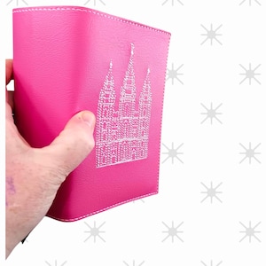 May include: A bright pink, leather-like cover with white stitching and a detailed white embroidered design of a building. The cover is held in a hand and appears to be a journal or notebook.
