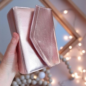 May include: A rose gold clutch with a textured finish. The clutch has a flap closure and a rectangular shape. The bag is held by a hand and is set against a blurred background with decorative lights and a mirror.