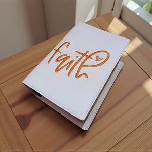 Op de afbeelding: Witte agenda met het woord "faith" geborduurd in oranje script, met een klein hartdetail. De agenda is open en toont de binnenpagina's. De kaft is gemaakt van een glad, leerachtig materiaal.