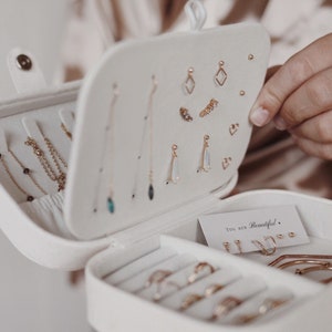 May include: White jewelry box with a velvet interior. The box is open and filled with gold earrings and rings. The box has a small card inside that says "You are Beautiful."