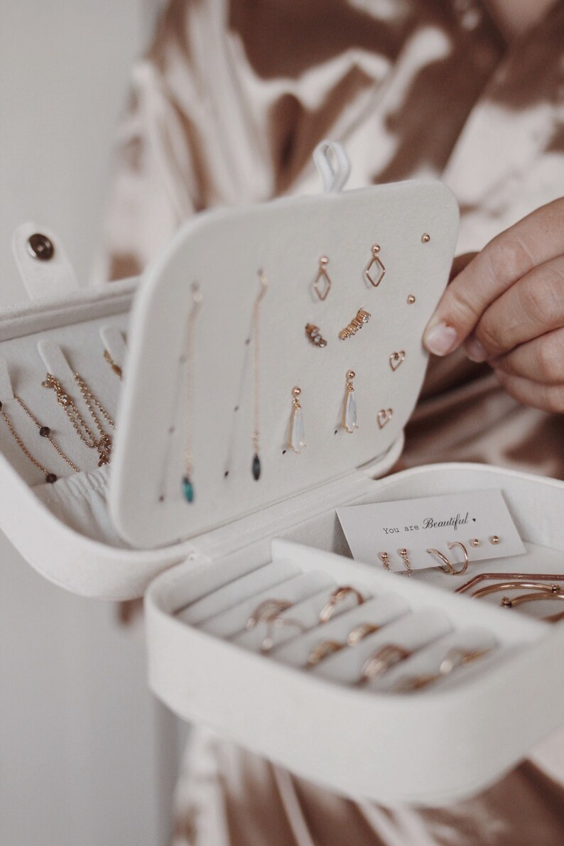 May include: White jewelry box with a velvet interior. The box is open and filled with gold earrings and rings. The box has a small card inside that says "You are Beautiful."