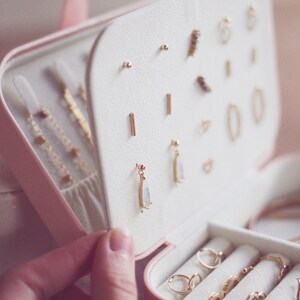 May include: A pink jewelry box with a white interior. The box is open and contains a variety of gold earrings and rings. The earrings are arranged in rows, while the rings are in a separate compartment. The box is made of a soft, velvety material.