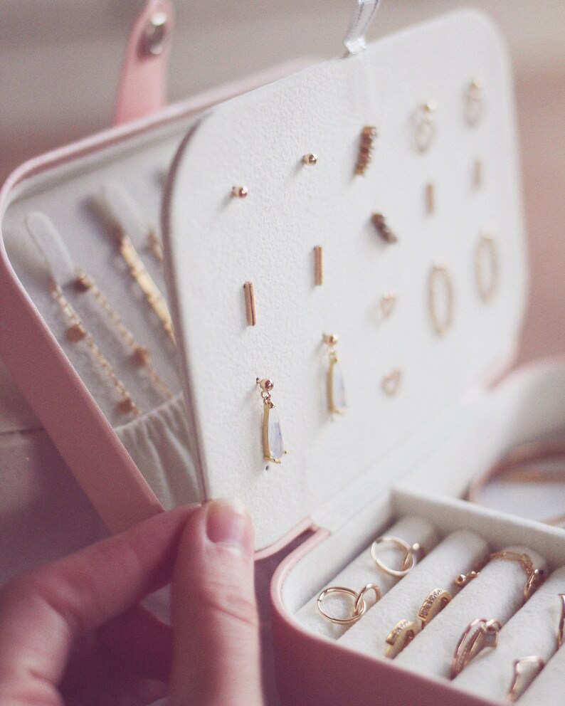 May include: A pink jewelry box with a white interior. The box is open and contains a variety of gold earrings and rings. The earrings are arranged in rows, while the rings are in a separate compartment. The box is made of a soft, velvety material.