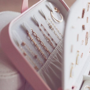 May include: A pink jewelry box with a white interior, open to reveal rows of gold chains and a gold ring. The box is lined with soft white fabric.