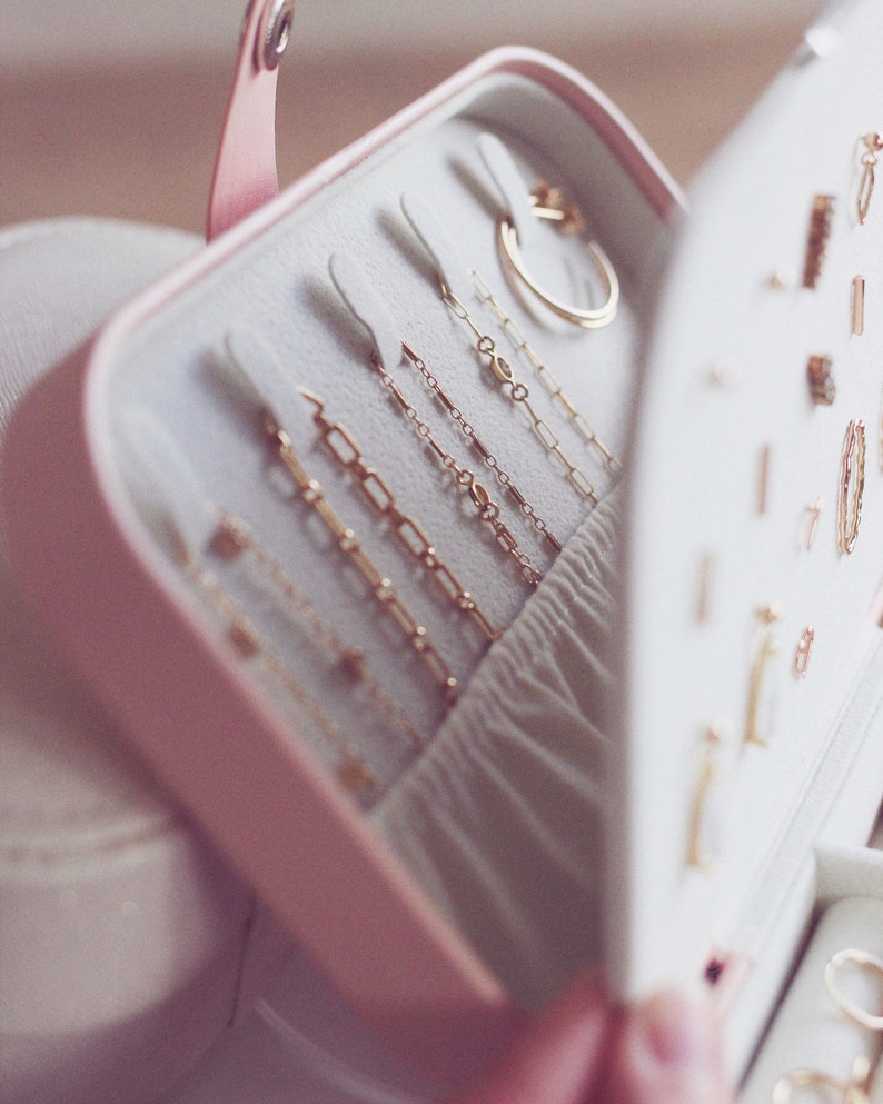 May include: A pink jewelry box with a white interior, open to reveal rows of gold chains and a gold ring. The box is lined with soft white fabric.