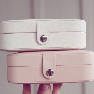 May include: Two stacked oval-shaped jewelry boxes. The top box is white with a fabric texture and a silver button closure. The bottom box is pink with a smooth finish and a silver button closure.