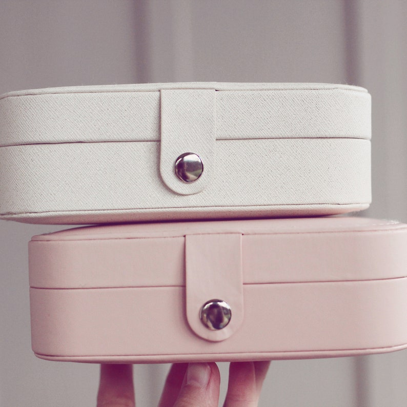 May include: Two stacked oval-shaped jewelry boxes. The top box is white with a fabric texture and a silver button closure. The bottom box is pink with a smooth finish and a silver button closure.