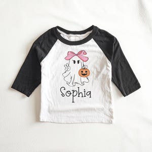 May include: White and black baseball tee shirt with a ghost wearing a pink bow and holding a pumpkin. The shirt says "Sophia" in black text.