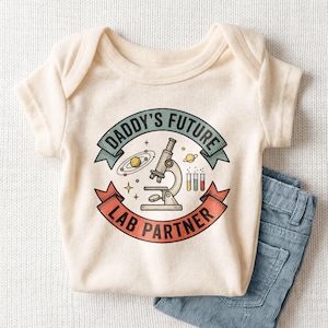 May include: Cream-colored baby bodysuit with the text "DADDY'S FUTURE LAB PARTNER" in a banner design. The graphic includes a microscope, planets, and test tubes. Light blue denim jeans are folded next to the bodysuit.