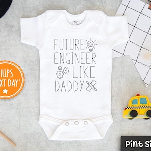 May include: White baby onesie with the text "FUTURE ENGINEER LIKE DADDY" in black. The onesie has a lightbulb, gears, and ruler graphic. A yellow toy taxi and other baby items are in the background.