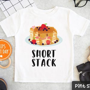 May include: White t-shirt with a graphic of a short stack of pancakes with strawberries and blueberries. The text "Short Stack" is printed below the pancakes.