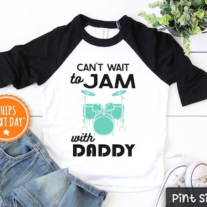May include: A white and black baseball tee shirt with a graphic of a drum set and the text "Can't wait to JAM with DADDY".