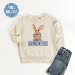May include: A beige sweatshirt with a brown bunny wearing sunglasses and a blue bow tie. The bunny is holding a blue and white checkered sign that says "Charlie".