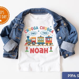 May include: White toddler t-shirt with a colorful train graphic and the text "CHUGGA CHUGGA TWO" above the train. The name "NOAH" is below the train. The shirt is paired with a blue denim jacket.