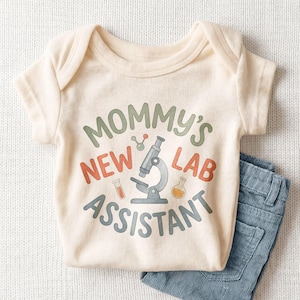 May include: A cream-colored baby onesie with the text "Mommy's New Lab Assistant" in green, orange, and gray. The design includes a microscope and lab equipment. Light blue corduroy pants are visible on the right side of the image.