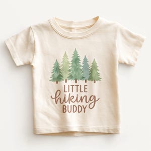 May include: Cream-colored toddler t-shirt with a graphic of five green pine trees and the words "LITTLE hiking BUDDY" in brown script. The shirt has short sleeves and a crew neck.