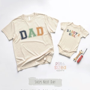 May include: Two beige t-shirts and a onesie. One t-shirt has the word "DAD" in navy, green, and orange. The onesie says "DADDY'S GIRL" in colorful letters. A wrench and screwdriver are next to the onesie.