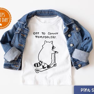 May include: White t-shirt with a black and white illustration of a raccoon on a scooter, and the text "OFF TO COMMIT TOMFOOLERY." A blue denim jacket is draped over the shirt. The shirt is labeled "Pint Sized."