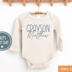 May include: A cream-colored long-sleeved bodysuit with the names "Grayson Matthew" printed in blue on the front.