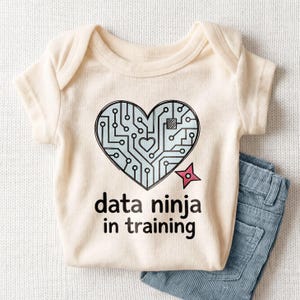 May include: Cream-colored baby bodysuit with a heart-shaped circuit board design and the text "data ninja in training." A pink ninja star is next to the heart. Light blue denim jeans are folded next to the bodysuit.