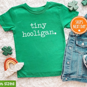 May include: Green short-sleeved t-shirt with the text "tiny hooligan*" in white. The shirt is laid out on a white fuzzy surface with a rainbow-colored yarn decoration and a denim jacket.