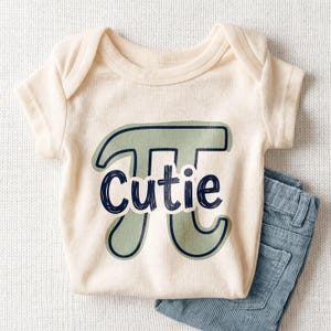 May include: Cream-colored baby onesie with a green and navy blue Pi symbol and the word "Cutie" in navy blue script. Light blue corduroy pants are folded next to the onesie. The outfit is displayed on a white, textured surface.