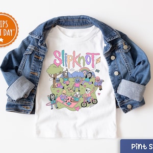 May include: White t-shirt with a colorful graphic featuring the word "Slipknot" and cartoon characters enjoying a picnic. The shirt is paired with a blue denim jacket. The image includes a "Ships Next Day" badge.