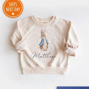 May include: Cream-colored sweatshirt featuring a watercolor illustration of Peter Rabbit wearing a blue jacket. The name "Matthew" is written in script below the image. The sleeves are cuffed. The sweatshirt is pint sized.
