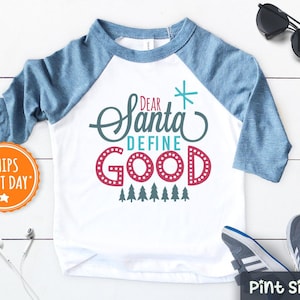 May include: A white and blue raglan t-shirt with the text "Dear Santa Define Good" in a festive font. The shirt features a red and white star and a row of green trees.