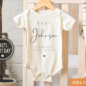 May include: A white baby onesie with black text that reads "Baby Johnson Coming Dec 2022".