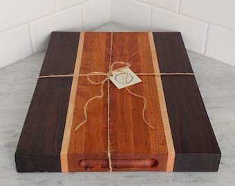 May include: A rectangular wooden cutting board with a dark brown border, a light brown stripe, and a reddish-brown center. The board is tied with twine and has a small tag. The cutting board has a handle cut out on one side.