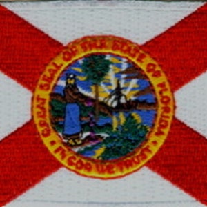May include: A red and white embroidered patch featuring the state seal of Florida. The seal depicts a sun, a tree, and a woman in a blue dress standing on a beach. The text "Great Seal of the State of Florida" and "In God We Trust" are inscribed around the seal.