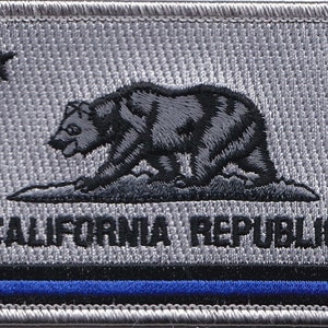 May include: Embroidered patch featuring the California state flag design. The patch has a silver background with a black bear, a star, and the words "CALIFORNIA REPUBLIC." A blue stripe is at the bottom.