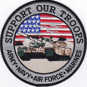 May include: A round, gray and black embroidered patch with the text "Support Our Troops" around the edge. The center of the patch features an American flag with a tank in front of it. The text "Army - Navy - Air Force - Marines" is below the tank.