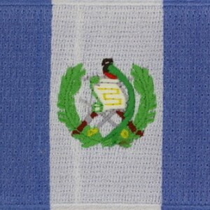 May include: A blue, white, and green embroidered patch featuring the national coat of arms of Guatemala. The coat of arms depicts a quetzal bird with a scroll in its beak, surrounded by a wreath of green leaves.