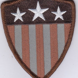 May include: Shield-shaped patch with a brown border. The top section is brown with three white stars. The lower section has alternating vertical stripes of light brown and gray. The patch is likely for a uniform or garment.
