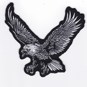 May include: Embroidered patch featuring a detailed, stylized eagle with outstretched wings. The eagle is depicted in shades of gray and white against a black background. The patch is designed to be sewn or ironed onto fabric.
