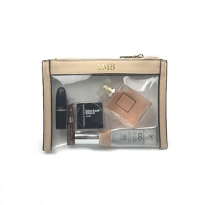 Könnte beinhalten: Eine beige, transparente Kosmetiktasche mit Reißverschluss. Die Tasche hat die Initialen "LMB" auf der Vorderseite aufgedruckt. Die Tasche enthält einen schwarzen Lippenstift, ein klares Parfümfläschchen, eine schwarze Make-up-Kompaktdose und eine Tube Foundation.