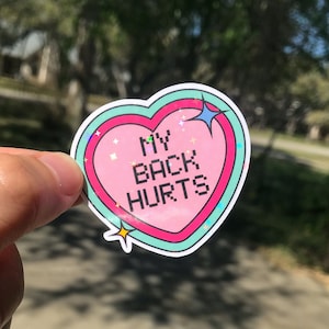 May include: A pink heart-shaped sticker with a glittery finish. The sticker has a blue outline and says "MY BACK HURTS" in white pixelated letters.