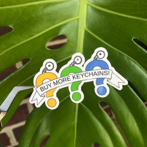 May include: Three colorful question mark keychains with a white banner that reads "BUY MORE KEYCHAINS!" The keychains are yellow, green, and blue.