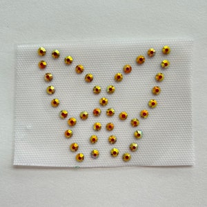May include: A gold butterfly design made of rhinestones on a white fabric background.