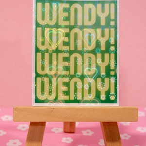 May include: A green holographic sticker with the text "WENDY!" repeated four times. The sticker has a rainbow iridescent effect and is surrounded by small hearts and circles.
