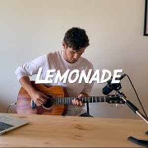 May include: A person playing an acoustic guitar with a microphone in front of them. The guitar is brown and has a white pickguard. The person is wearing a white t-shirt. The word "LEMONADE" is written in white letters on a black background.