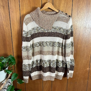 May include: A striped sweater in shades of brown, beige, and olive green hangs on a wooden hanger. The sweater features a cowl neck and button details on the shoulder. The horizontal stripes create a textured look.