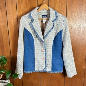 May include: A vintage blazer with a tweed-like fabric in light blue, beige, and white. The blazer features denim-like blue panels on the front and fringe detailing along the collar and hem. It has pink buttons and the brand name "Lorenza" on the label.