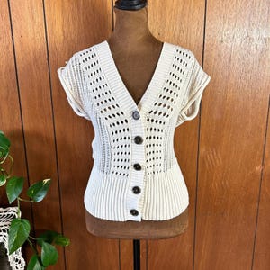 May include: Cream-colored short-sleeved cardigan with a V-neck and button closure. The knitwear features a mix of solid and openwork patterns. The cardigan is displayed on a mannequin against a wooden background.