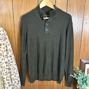 May include: A dark olive green sweater with a mock neck and three buttons. The long-sleeved sweater is hanging on a wooden hanger against a wooden background. The sweater has a ribbed hem and cuffs.