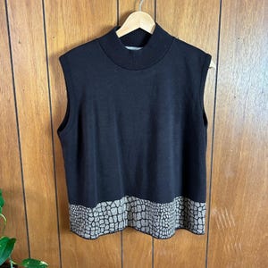 May include: A sleeveless black sweater with a mock turtleneck collar. The bottom hem features a textured, reptile-like pattern in shades of beige and gray. The sweater is hanging on a wooden hanger.