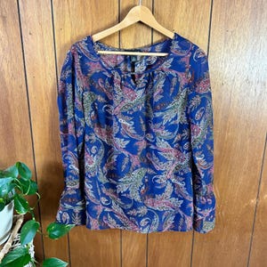 May include: A long-sleeved blouse in a vibrant blue hue, adorned with a paisley pattern in shades of pink, green, and white. The blouse features a keyhole neckline and bell sleeves, hanging on a wooden hanger.