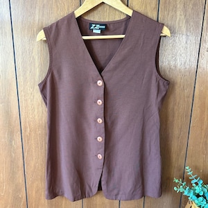 May include: A sleeveless brown vest with a V-neckline and six round, light brown buttons down the front. The vest is hanging on a wooden hanger against a wooden panel background. The vest has a small label near the neckline.